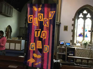 Banner of irregular vertical blue and mauve stripes with letters on orange squares spelling 'Glory to God'