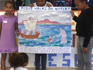 Children with a banner of a lake with a boat and Jesus standing on the water and lifting a sinking disciple