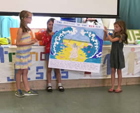 Two girls hold up a picture of waves curling over dry land as Moses walks across