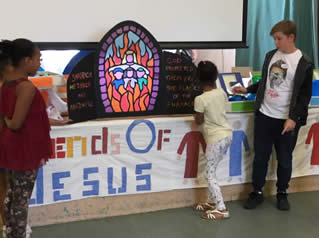 Children inspect a model of a stained glass window of 3 people surrounded by fire and embraced by a fourth figure