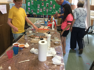 Two children and two helpers standing by a table on which are paint pots and partly-made bits of Roman armour