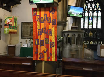 Banner of irregular vertical stripes of orange and gold and letters on dark squares spelling 'Praised the Lord'